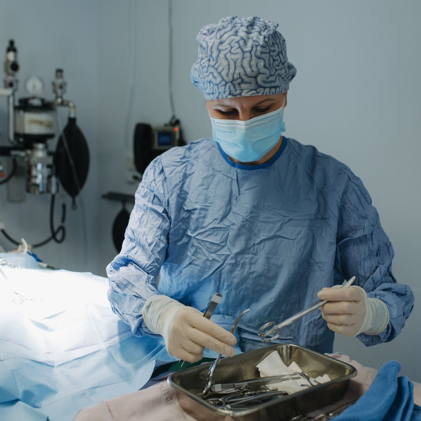 A person in surgical scrubs and a mask holds surgical instruments over a tray of tools in an operating room.